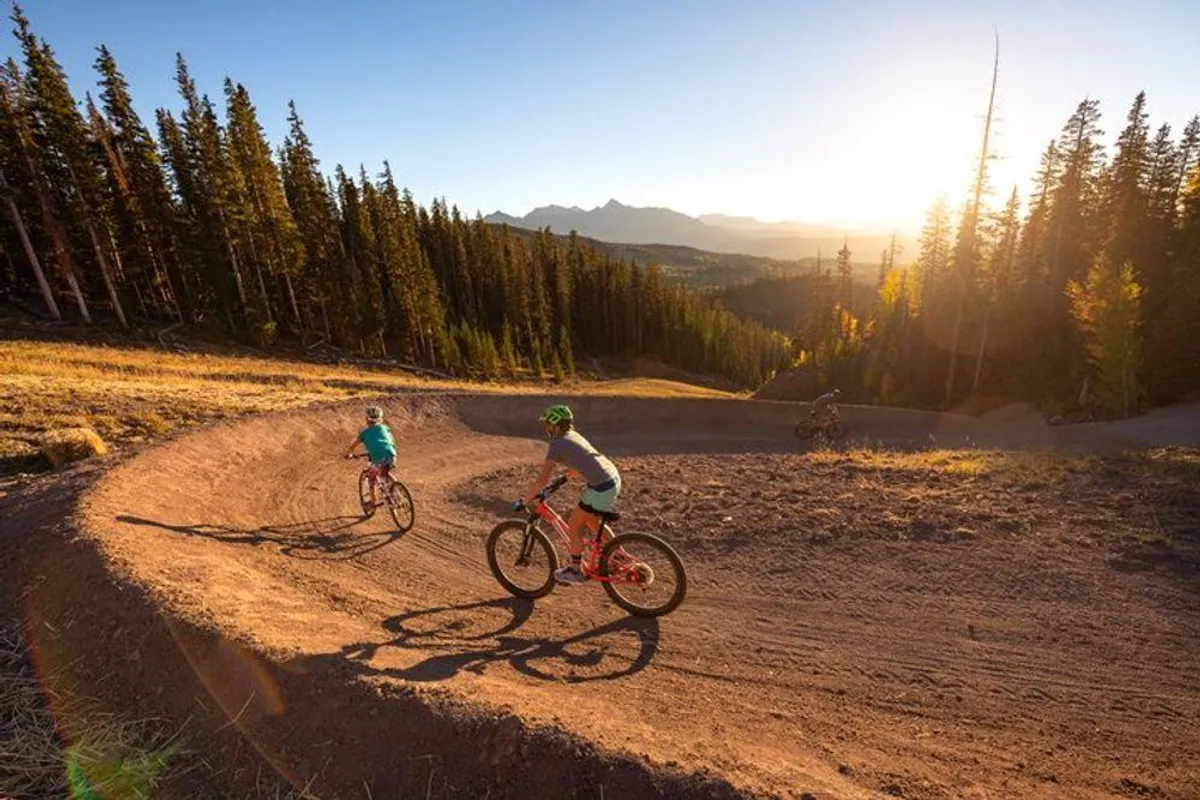 A group of mountain bikers navigate a winding trail through dense trees with majestic mountains in the distance. The golden light hints at late summer or early fall, highlighting the dynamic outdoor activities available.