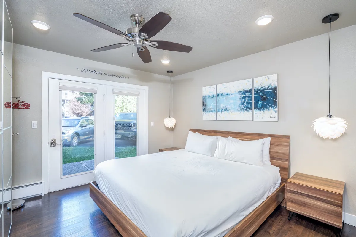 A softly lit bedroom featuring a comfortable king bed dressed in crisp, white linens. The room is awash in natural light from large windows, accentuated by soothing, neutral tones and calming artwork above the bed. Blackout curtains frame the windows, and a ceiling fan adds a touch of comfort. The space feels tranquil and ideal for couples or solo travelers seeking rest.