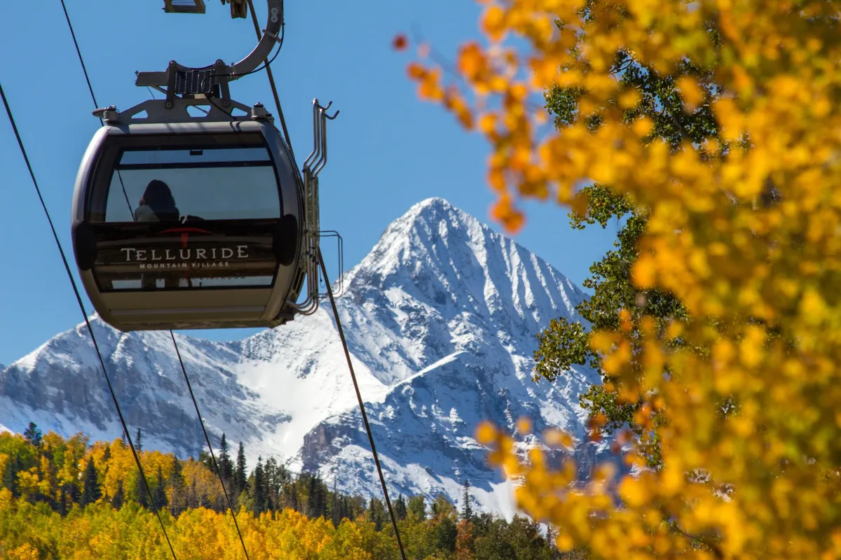 This vibrant scene features the Telluride gondola against a backdrop of snow-capped mountains and colorful autumn foliage. The gondola is prominently visible, emphasizing easy access to adventure, festivals, and the heart of town.