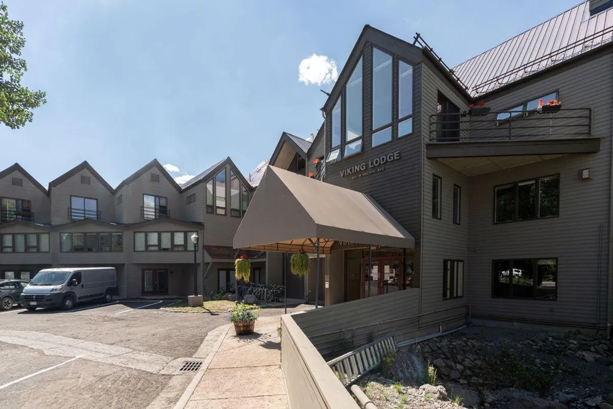 The image captures the welcoming exterior of the Viking Lodge, showcasing its entrance awning, inviting sign, and landscaped surroundings. Adjacent buildings and outdoor patio seating hint at the communal atmosphere and the lodge's central, walkable location.