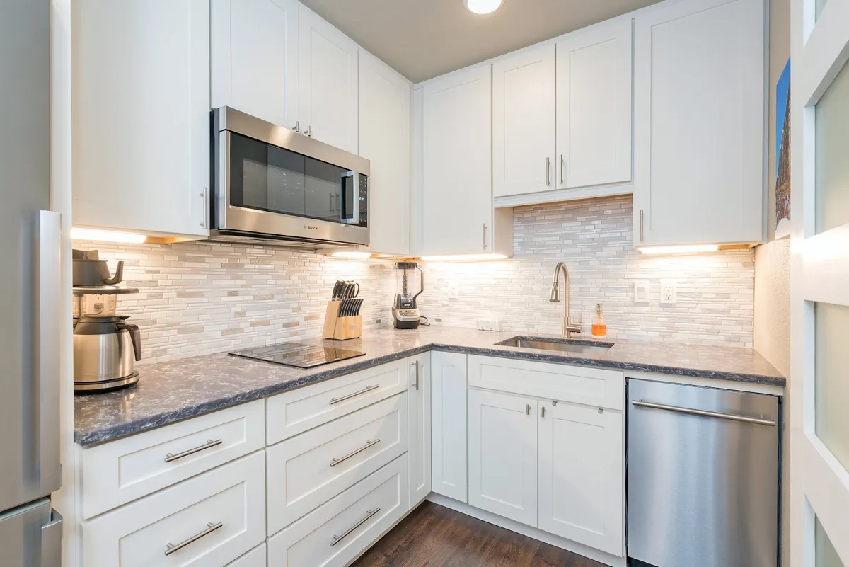 This image features a bright, inviting kitchen with a central island, stainless steel appliances, modern cabinetry, and a stylish tile backsplash. The space is well-lit and equipped for both cooking and socializing, showing kitchen utensils and seating around the island.