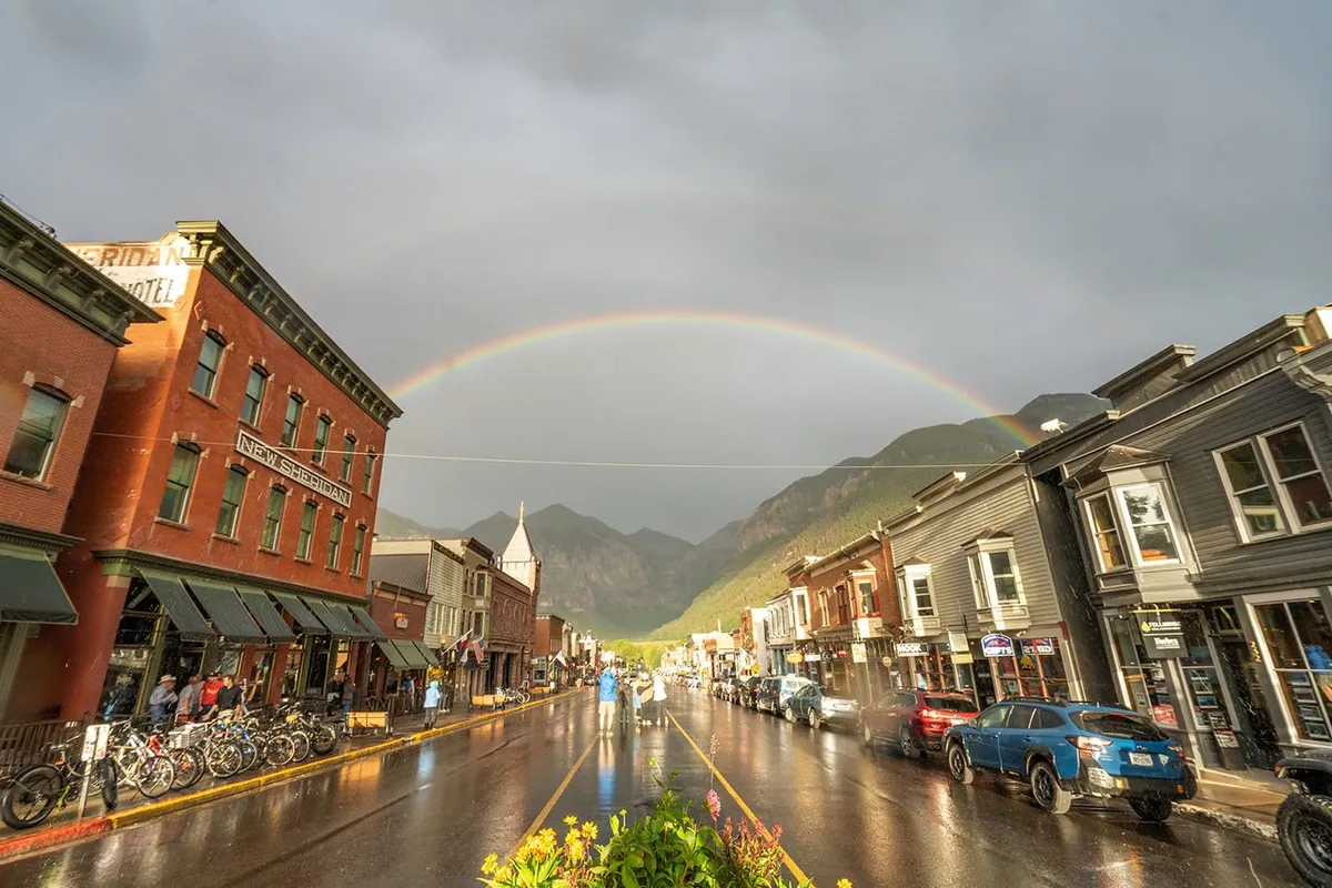 A vibrant Telluride street scene after a rainstorm, featuring historic buildings, bicycles, a rainbow overhead, and wet reflections on the street. The backdrop of mountains and the presence of outdoor cafes and shops underscore the town's walkability and charm.