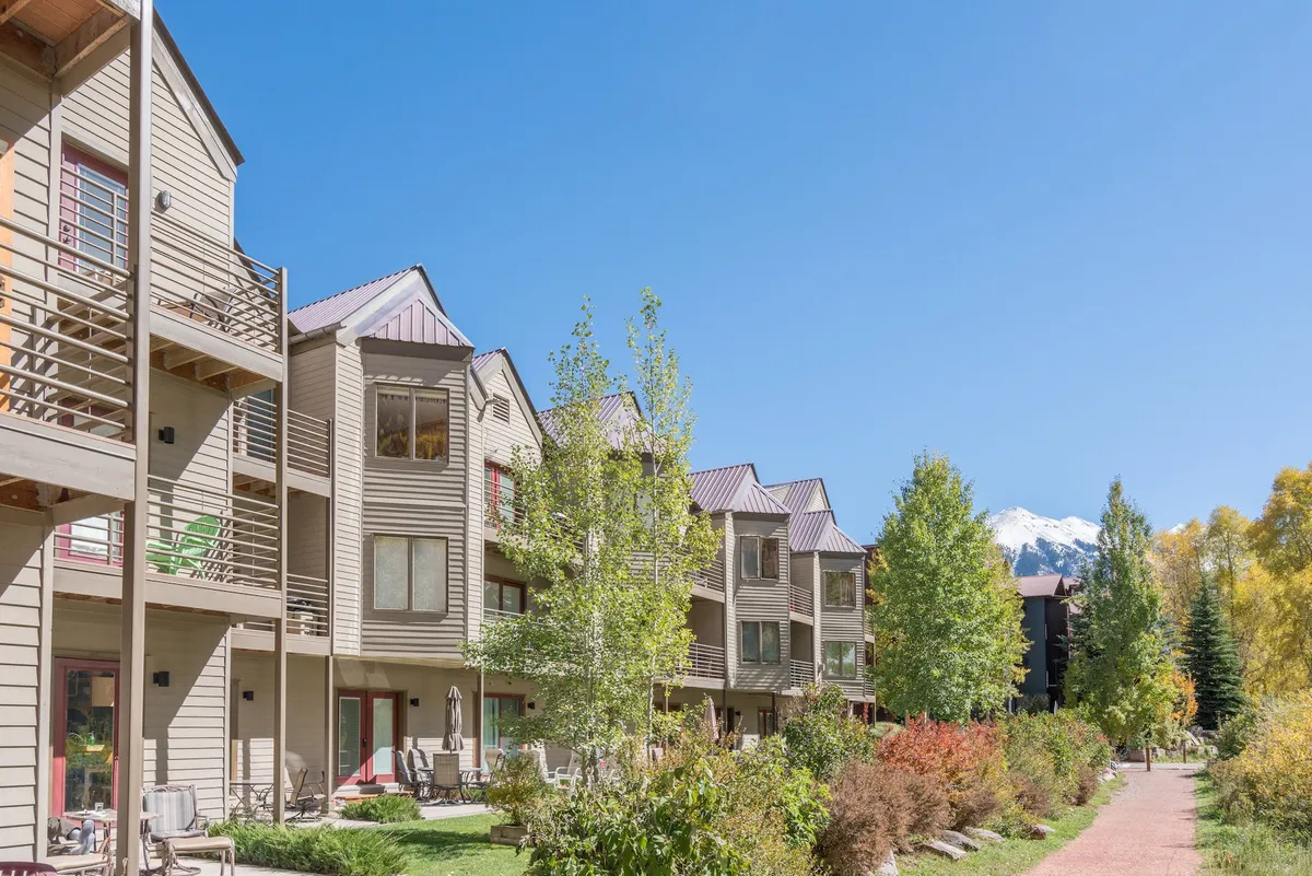 A row of buildings lines a tranquil pathway bordered by trees in full autumn color, with mountains rising in the background. The pathway is ideal for easy walks or stroller outings, while balconies suggest opportunities to relax and observe the scenery.
