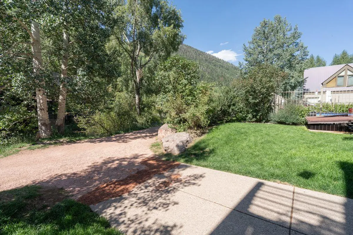 A sunlit pathway bordered by vibrant green grass and mature trees leads toward a stunning backdrop of mountains. The open lawn is ideal for picnicking, strolls, or enjoying nature together. The inviting, family-friendly green space is perfect for children to play or explore safely.