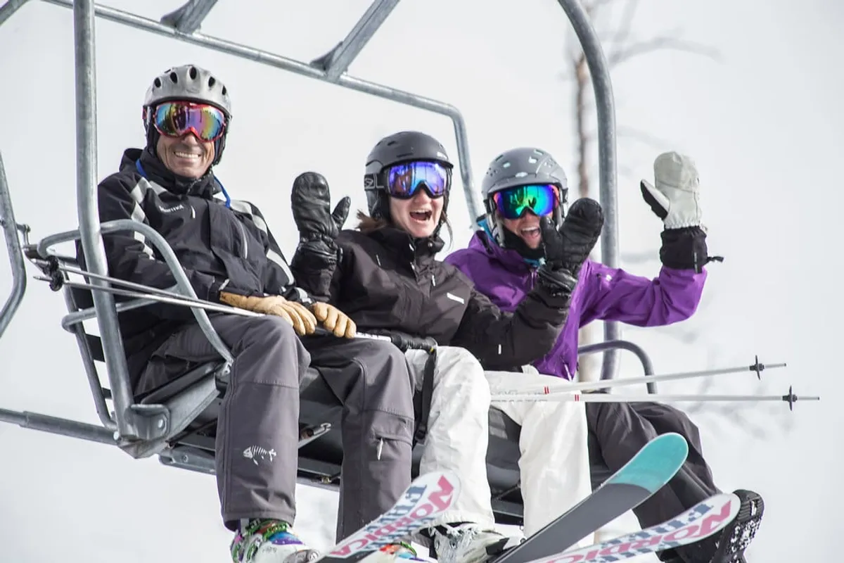 Three people are riding a ski lift above a snowy landscape, dressed in ski gear and ready to hit the slopes. The background features pristine snow, pine trees, and crisp mountain air, capturing the energy of Telluride's winter sports scene.