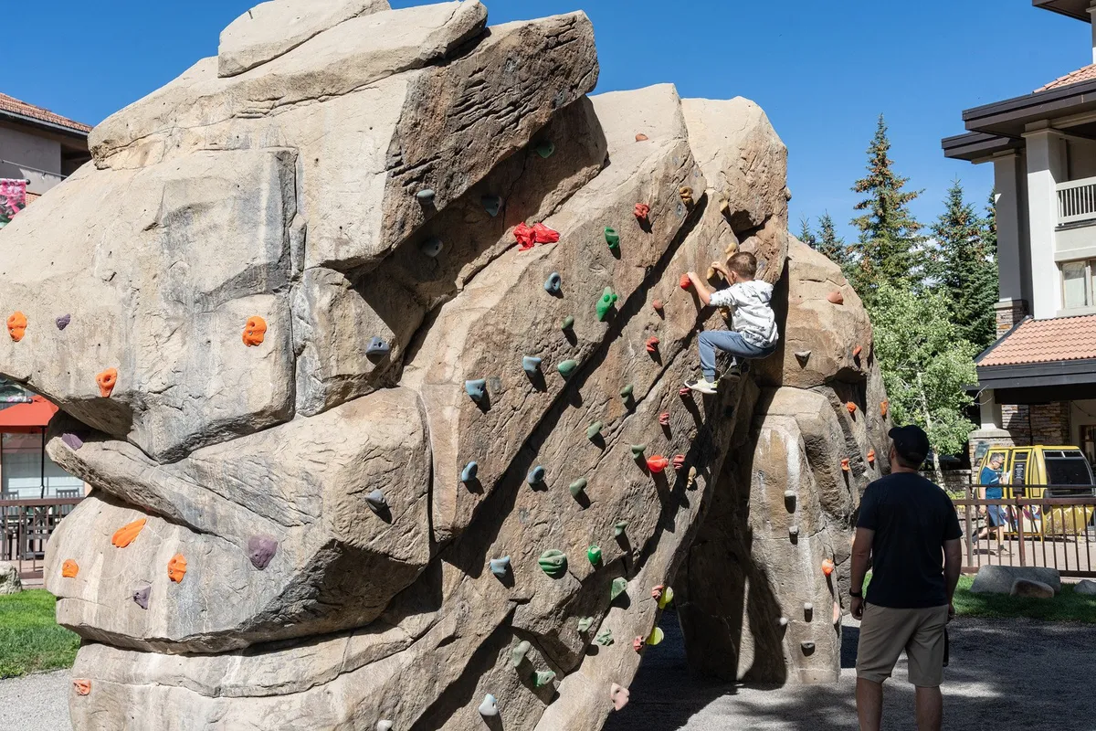 A child is climbing on an outdoor wall, closely watched by an adult, amidst a safe, playful setting. The scene radiates energy, family bonding, and the joy of outdoor activity for kids, set against a backdrop of greenery.