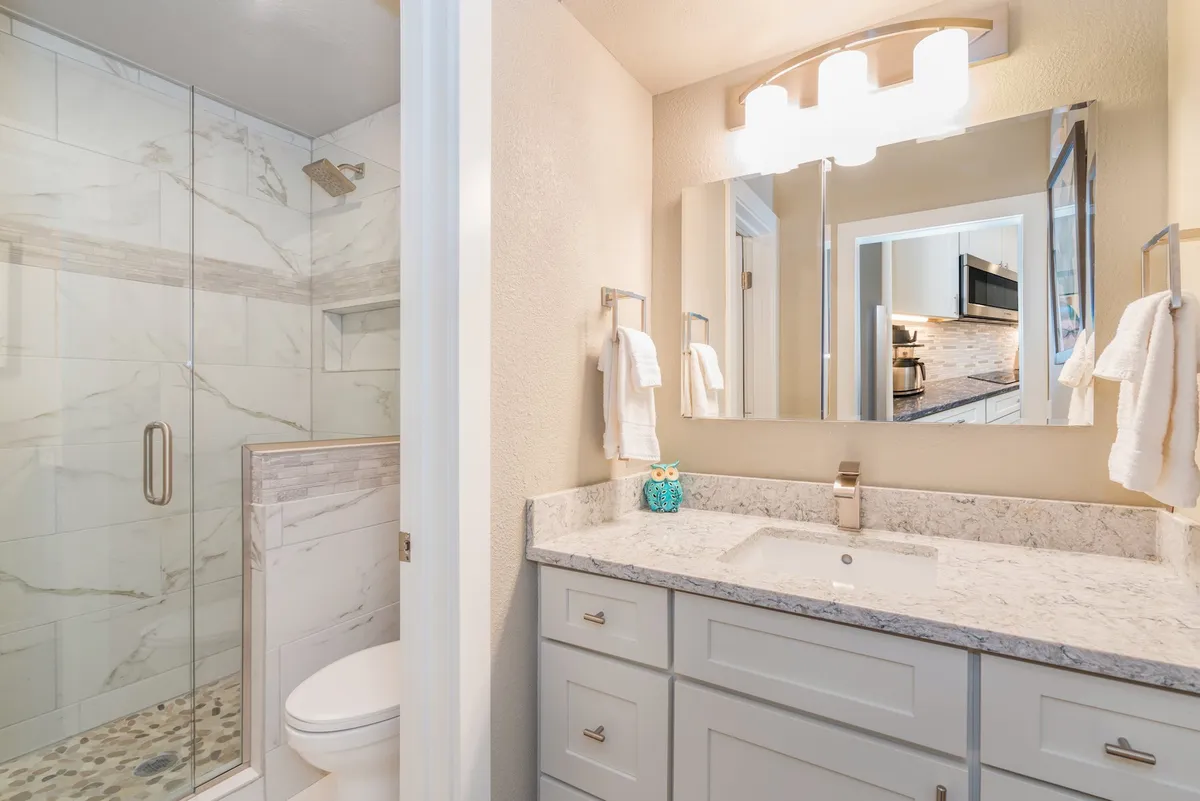 A modern, pristine bathroom featuring a glass-enclosed shower, marble countertop, and contemporary lighting. Decorative accents add warmth, while plush towels are neatly arranged, evoking a spa-like feel. The space feels fresh, inviting, and immaculately clean.