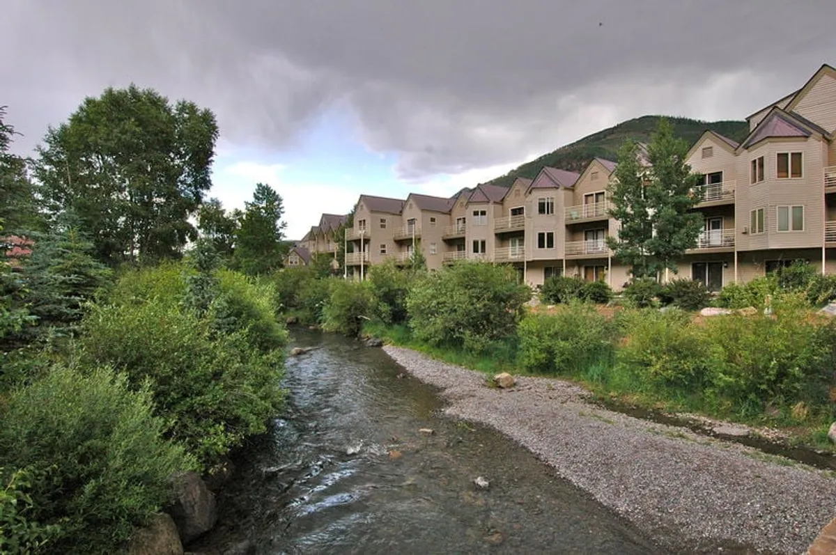 A peaceful stream runs alongside the building with mountains in the background. Trees line the riverbank, and the pathway invites morning walks or evening strolls along the River Trail.