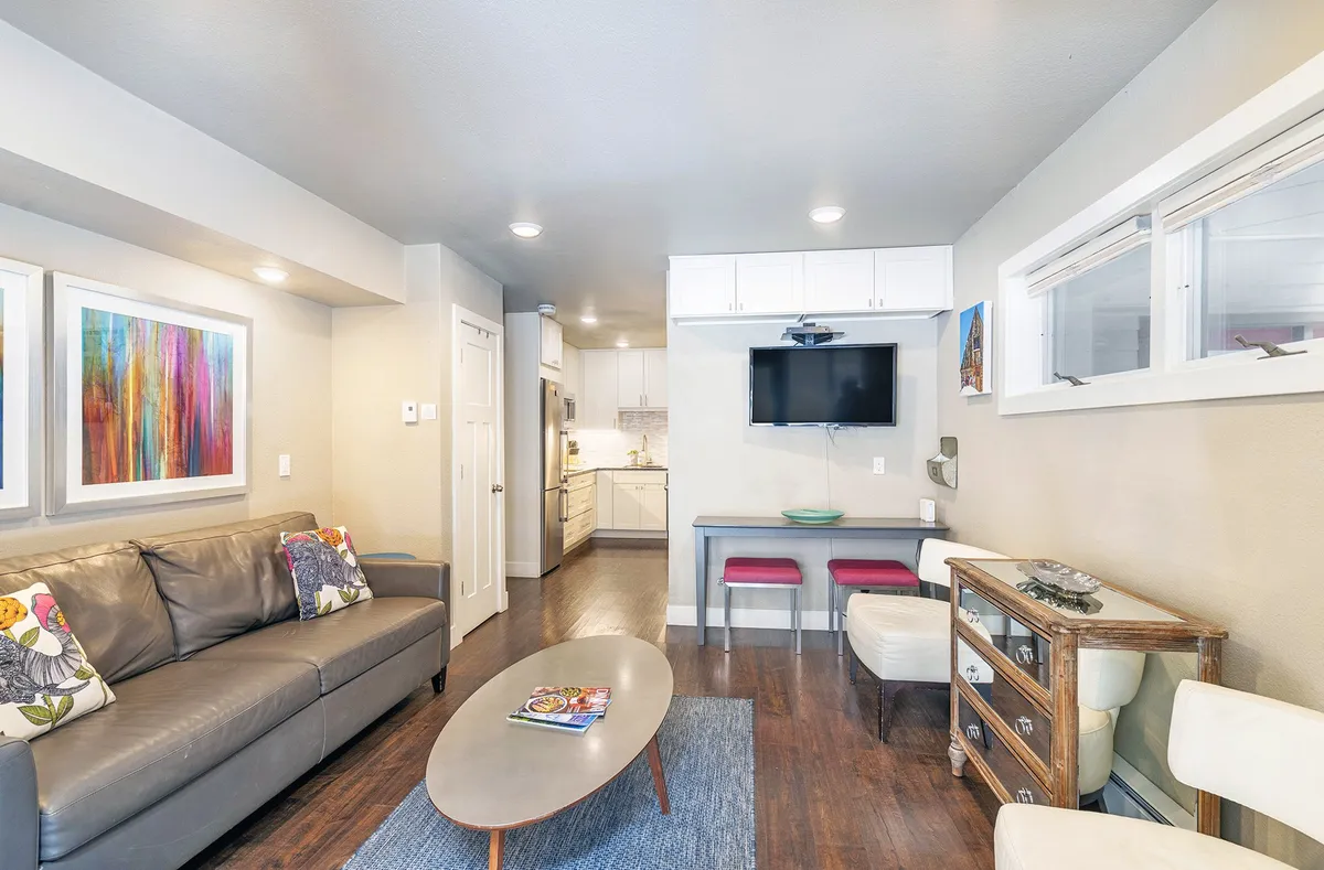 A stylish, modern condo living area featuring a plush sofa, inviting coffee table, sleek open kitchen, and vibrant artwork, all bathed in natural light. The space feels cozy yet contemporary, perfect for relaxing evenings or intimate meals.